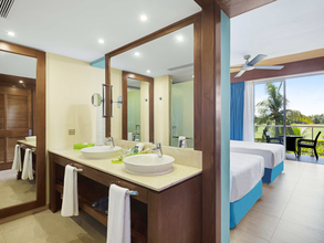 Bathroom with two sinks in front of a room with two single beds in front of a balcony with a table and chairs giving out on a palm tree and grass.